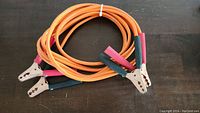 Photo of a pair of orange booster cables with metal clamps and insulated grips laid coiled on a dark surface.