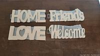 Four white wooden wall signs spelling HOME, Friends, LOVE, and Welcome arranged on a wooden surface.