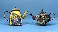 Two Chinese enameled mini teapots arranged side by side on a plain blue background, showing front and slightly angled views.
