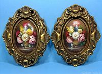 Pair of antique oval oil paintings on board depicting colorful floral bouquets in elaborate gold-toned frames against blue background.
