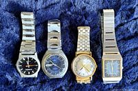Four vintage men's watches with various dial colors and metal bands displayed on a blue fabric surface.
