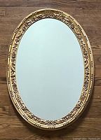 Front view of the entire vintage oval mirror showing the detailed ornate gilt frame and clean mirror glass lying flat on wood floor.