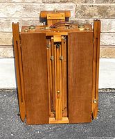 Front view of folded wooden easel showing the handle, folding panels, and brass hardware.