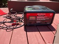 Black Motomaster manual battery charger with cables and clamps on red wooden surface, front view showing label and analog gauge.