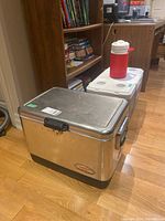 Photo showing the set of three coolers arranged on a hardwood floor near a bookshelf: large silver metal Coleman chest cooler, white plastic chest cooler, and red drink cooler jug on top.