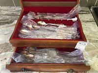 Top view showing various forks, spoons, and knives wrapped in plastic bags and arranged in a wooden cutlery chest with red felt compartments.