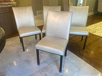 Four matching dining chairs arranged with two in front and two behind, showing light beige vinyl upholstery and black metal legs.