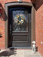 Front door with decorative wreath hung on glass panel and ceramic dog statue sitting on the doorstep near the brick wall.