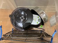 Photo showing the T-Fal Actifry fryer with transparent lid, insert, control panel, and metal Kaiser cake pan on the side plus metal grids inside a clear plastic bin.