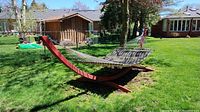 Side view of hammock on red curved wooden stand on green grass with a fence and houses in the background.