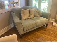 Side angled view of the green upholstered two-seat love seat with three throw pillows and four wooden legs on wood floor by windows.