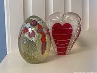 Two glass paperweights placed side by side on a white surface, showing egg-shaped Murano and heart-shaped Chinese paperweight