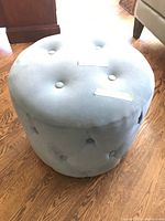 Front-right angled view of light gray round tufted ottoman showing button tufting and fabric upholstery on wood floor.