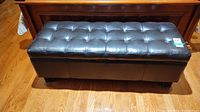 Front view of black tufted faux leather storage ottoman on wooden floor.