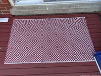 Full view of the rectangular outdoor rug with red and white geometric diamond pattern placed on wooden porch floor, showing overall condition and pattern detail.