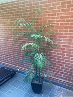 Full view of artificial plastic palm tree about 89 inches high against a brick wall, potted in a black square pot on tiled floor.
