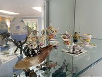 Porcelain miniatures displayed in glass cabinet including two miniature tea sets, ceramic egg on pedestal, snail seashell figurine, and other small porcelain items.