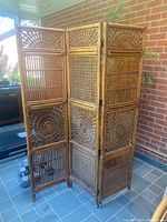 Full view of the three-panel bamboo room divider unfolded, showing woven sunburst motifs and vertical bamboo slats in warm natural tones against a brick wall background.