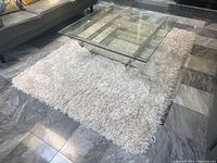 White shag pile area rug with thick plush texture under transparent glass coffee table on tiled floor, showing size and condition.