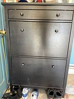 Front view of black shoe storage cabinet showing two tilt-out racks and one top drawer. Visible scratches on surface.