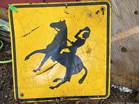 Front view of the metal horse crossing sign showing horse and rider silhouette on yellow background with black border. Visible cracks and rust indicate weathered outdoor condition.