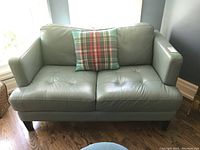 Grey leather loveseat sofa with two cushions and plaid throw pillow in front of window on wooden floor.