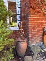 Side angle view showing the metal planter with metal flower decor inside, outdoors by brick wall and greenery.