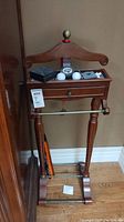 Front view of wood valet stand showing drawer, curved hanger with brass finial, and various items on top (not included).