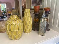 Photo showing two yellow West Elm terracotta vases with geometric pattern, pair of brown candlesticks with orange round accent pieces, and a rectangular decorative glass bottle with metallic details and spherical stopper.