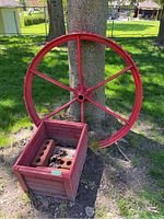 Metal farm wheel leaning against a tree outdoors with a red wooden box in front filled with bricks and debris.