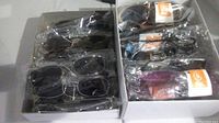 Two boxes of assorted new sunglasses with rectangular and oval dark lenses, various frame colors including black and purple, some individually wrapped in plastic.