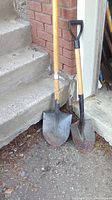 Two shovels leaning against a wall near stairs, showing full length and condition