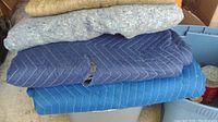 Top view showing stacked moving blankets and pads including blue quilted blankets and beige carpet-like pad.