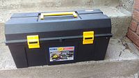 Closed view of the 22 inch black plastic toolbox with yellow latches and handle placed outdoors on concrete steps.