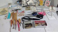 Wide shot of multiple sewing tools including scissors, buttons in tins, wooden handled tools, measuring devices, pins and needles spread out on a table.