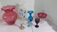 Group of seven colored and art glass pieces including vases, glasses, and a pedestal bowl arranged on a white surface.