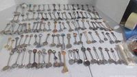 Wide shot showing about 105 souvenir spoons laid out on a white surface in rows, displaying the varied sizes and handle designs.