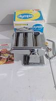 Olympia pasta maker next to its original box, showing its metal build and manual crank handle.