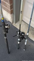 Three fishing rods with reels standing on pavement near a garage door, showing different designs and colors.