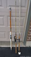 Three vintage fishing rods standing vertically against a garage door, showing rod coloration, handles and condition.