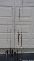 Six fishing rods standing upright showing various colors and handles against garage door