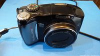 Front angle view showing black Canon PowerShot SX100 IS camera with 10x optical zoom and mode dial.