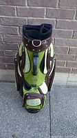 Front view of green and brown ladies golf bag with cream patterned pockets, standing upright against brick wall.