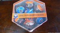 Top view of 7 visible Lionel Trains ornaments inside a sealed hexagonal collectible box showing blue and orange colors and train graphics.