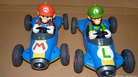 Front angle view of Mario and Luigi toy cars side by side, showing detailed character figures with steering wheels and blue car bodies.
