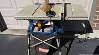 Front angle view of RYOBI table saw showing blade, controls, dust bag, and folding stand.