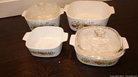 Four CorningWare L'Echalote Spice of Life vintage casserole dishes, three with handles and two with glass lids, all white ceramic with vegetable decoration along sides.