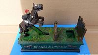 Top and side view of Design Toscano cast iron mechanical bank showing horse and rider jumping over gate on green base with embossed DETAIL.