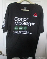 Full view of the black Conor McGregor UFC T-shirt hanging on a hanger, showing front graphics.