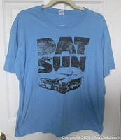 Light blue T-shirt hanging on a hanger, front view showing large black distressed print of the text 'DATSUN' with an illustrated image of a vintage 1976 Datsun B210 car below the text.